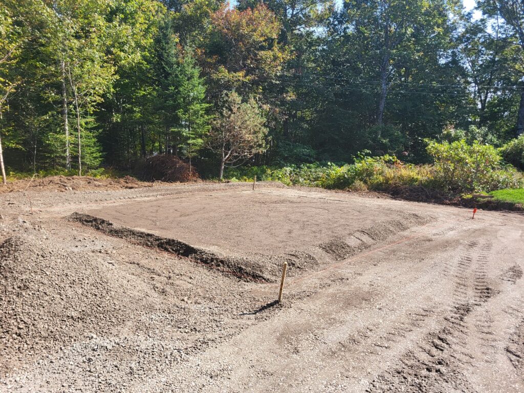 A wide view of a prepared building pad consisting of compacted brown granular fill. The rectangular pad features distinct, sloped edges and sits within a cleared area bordered by a dense forest. Orange string lines and wooden stakes mark the perimeter for the upcoming foundation work.